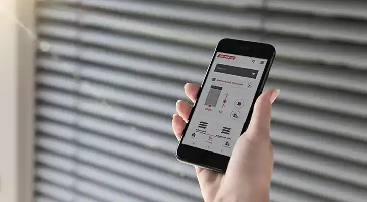 Person hält Smartphone mit Steuerungs-App vor Jalousien